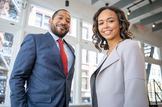 A man and woman both in business suits are standing looking towards the viewer. Both people are smiling. They are standing in a large windowed office.