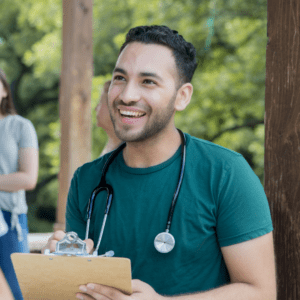 A smiling doctor in green scrubs holding a clipboard. The doctor is a young man with dark short hair.