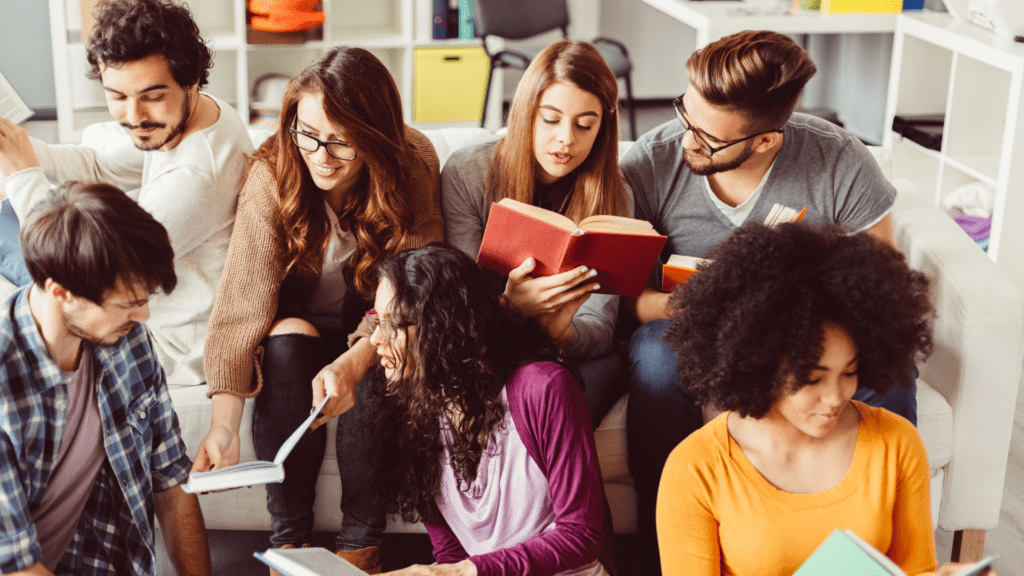 A group of young adults sitting together on a sofa in an office setting. They are all reading books. A couple of the people are showing their books to the others.